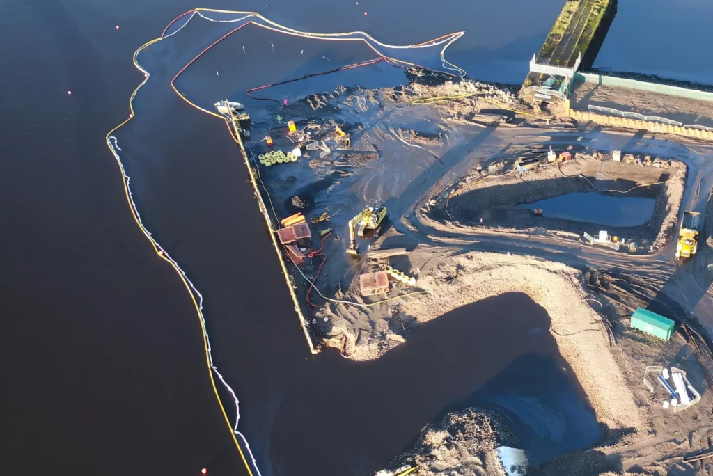 Aerial view of oil containment booms deployed around a shoreline construction site to prevent oil and debris from spreading into surrounding water during environmental cleanup operations.